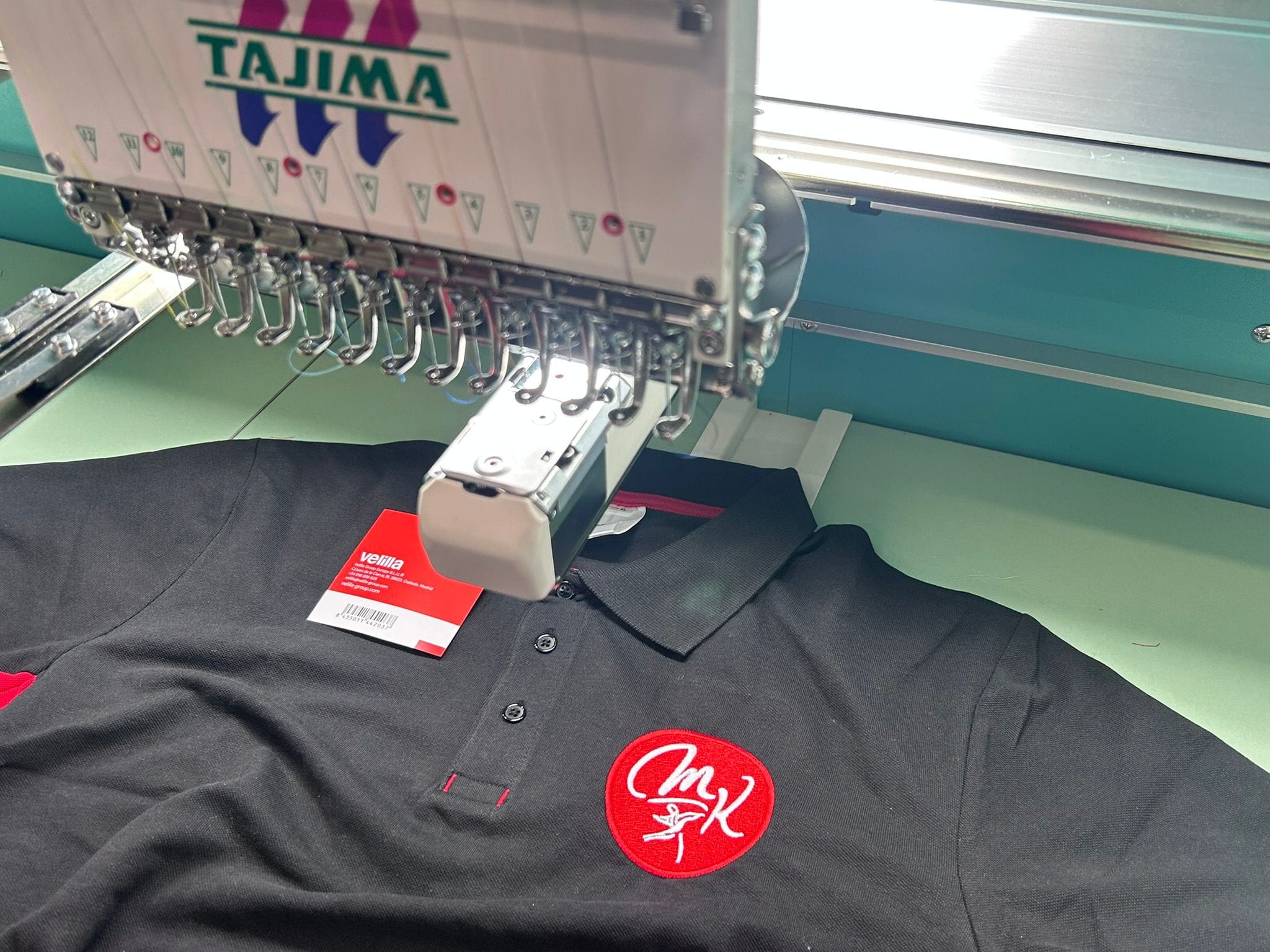 Polo noir avec un écusson brodé rond rouge et blanc, sous la tête de la machine à broder