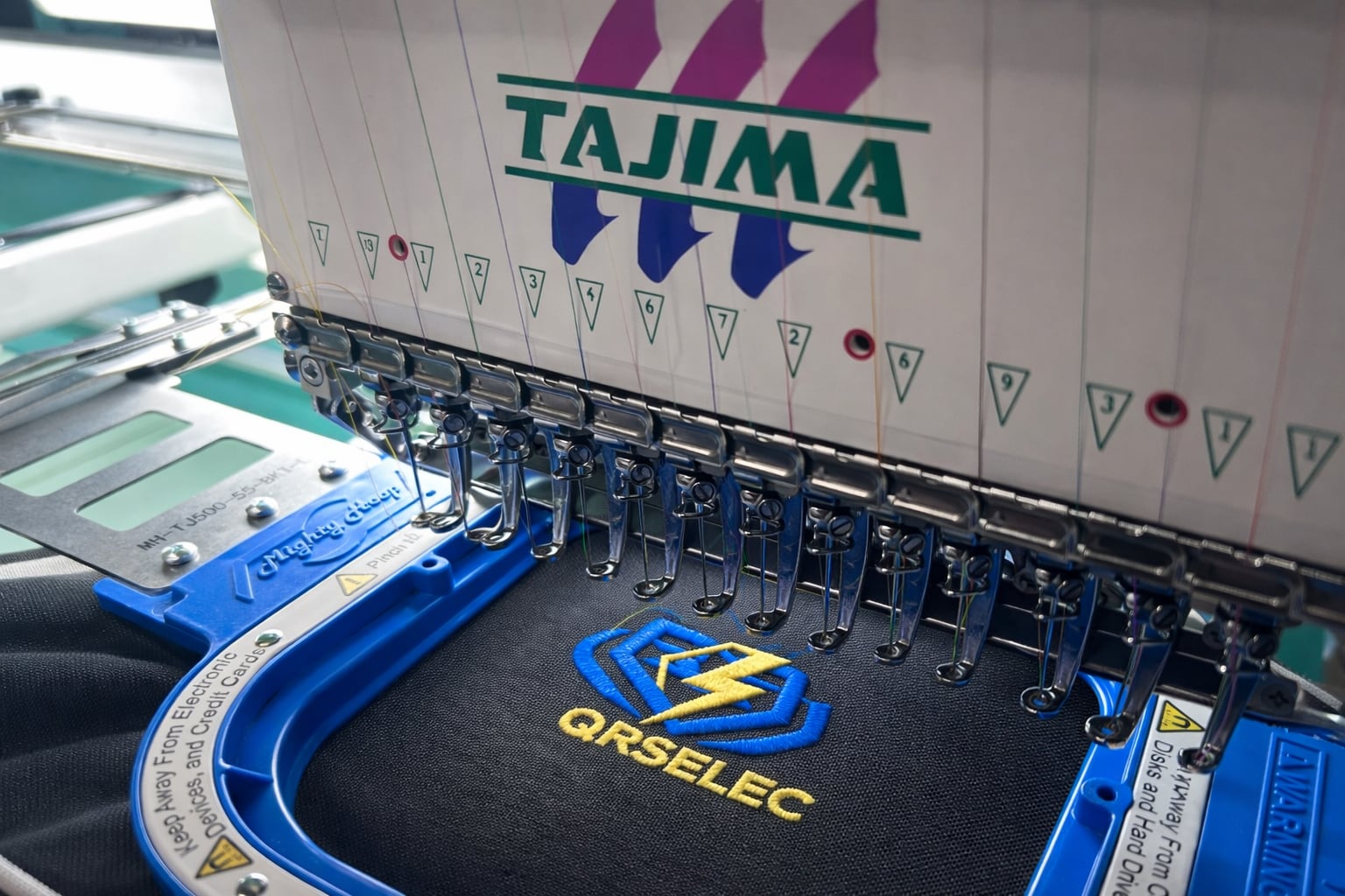 Machine à broder Tajima en train de broder le logo QRSELEC bleu et jaune sur un textile noir